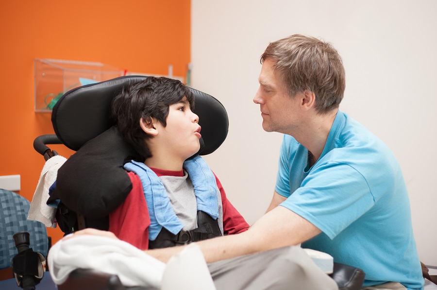 young boy with medical complexity, sitting in a power wheelchair inside a school or medical facility, facing a young man crouched down to assist.